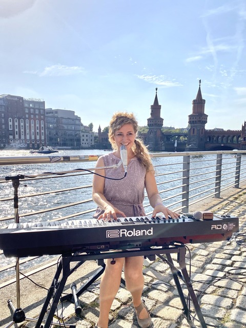 Eine Sängerin steht an einem Keyboard und singt. Im Hintergrund ist Wasser zu sehen.